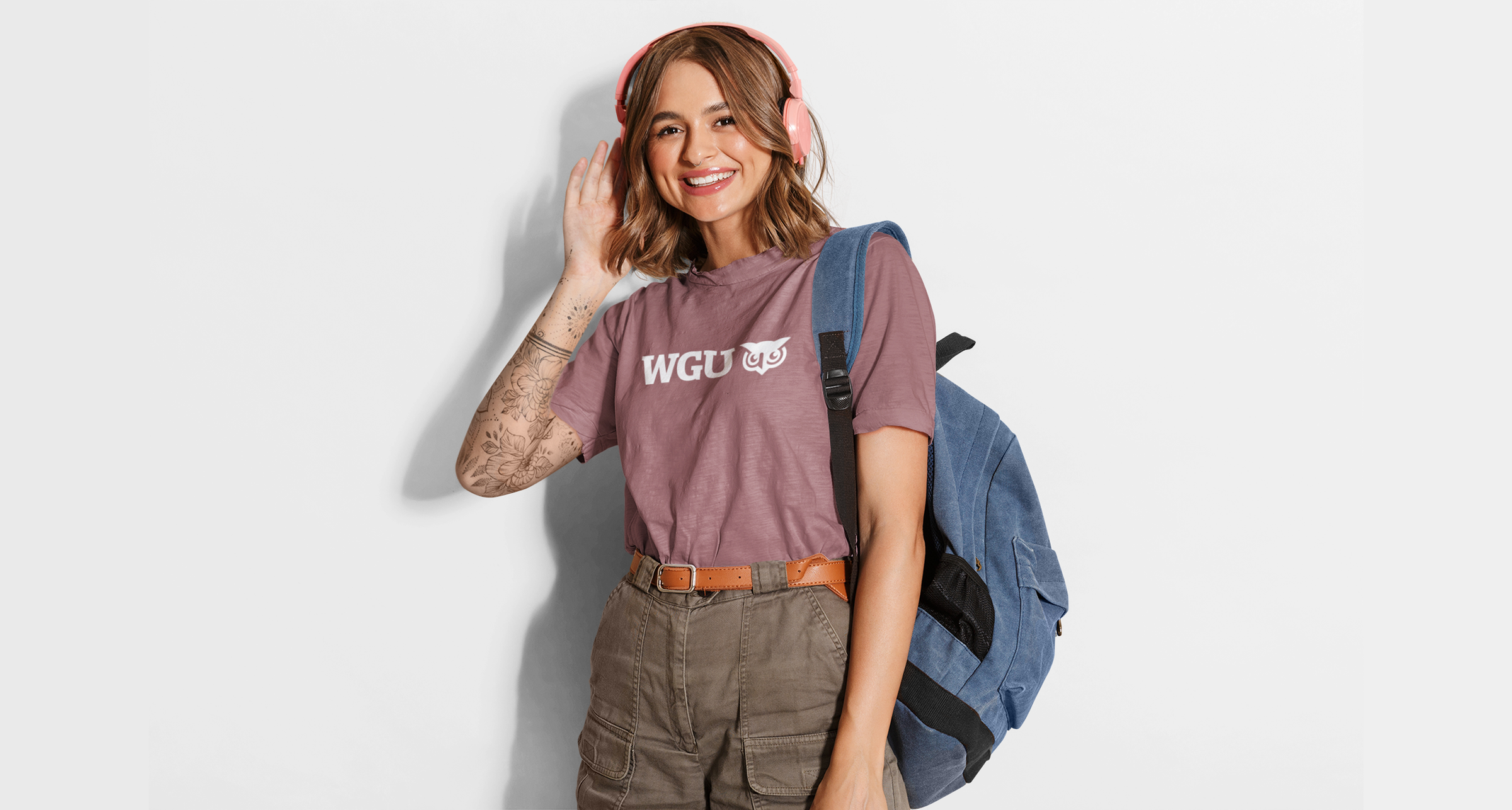 Girl in mauve shirt with WGU decoration wearing a blue backpack and pink headphones in front of cream background